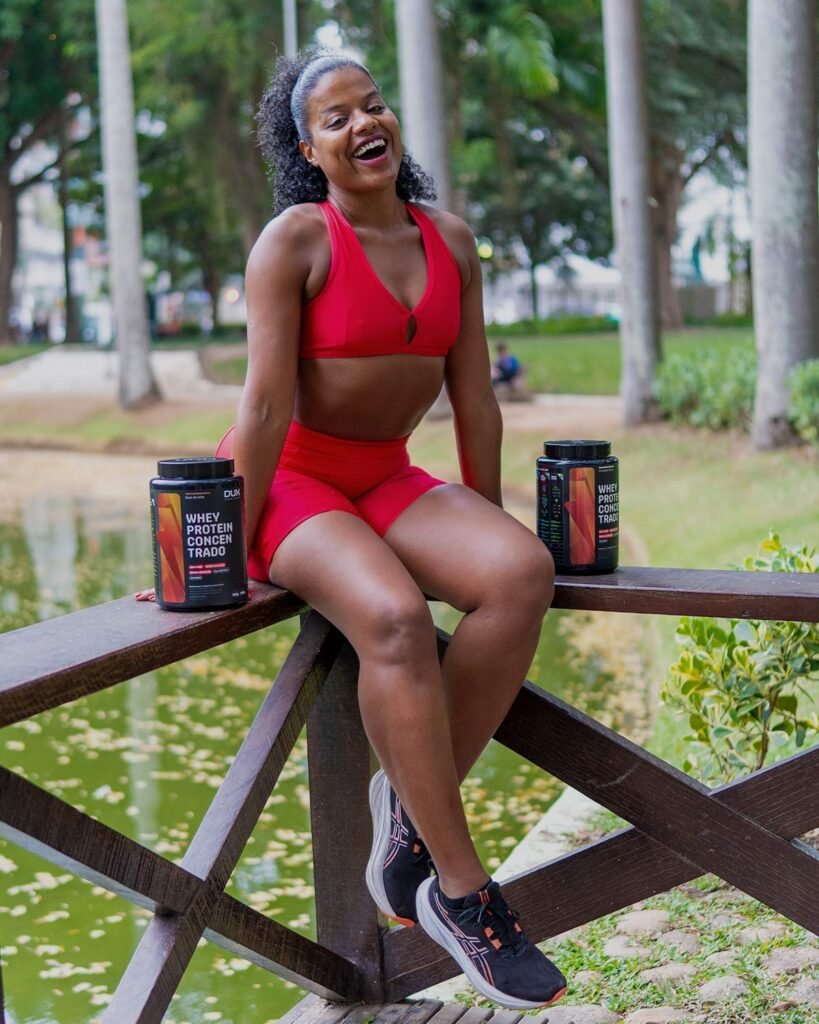 Luana Santinni em momento pós-treino ao ar livre, sentada em ponte de parque com suplementos de whey protein após atividade física.